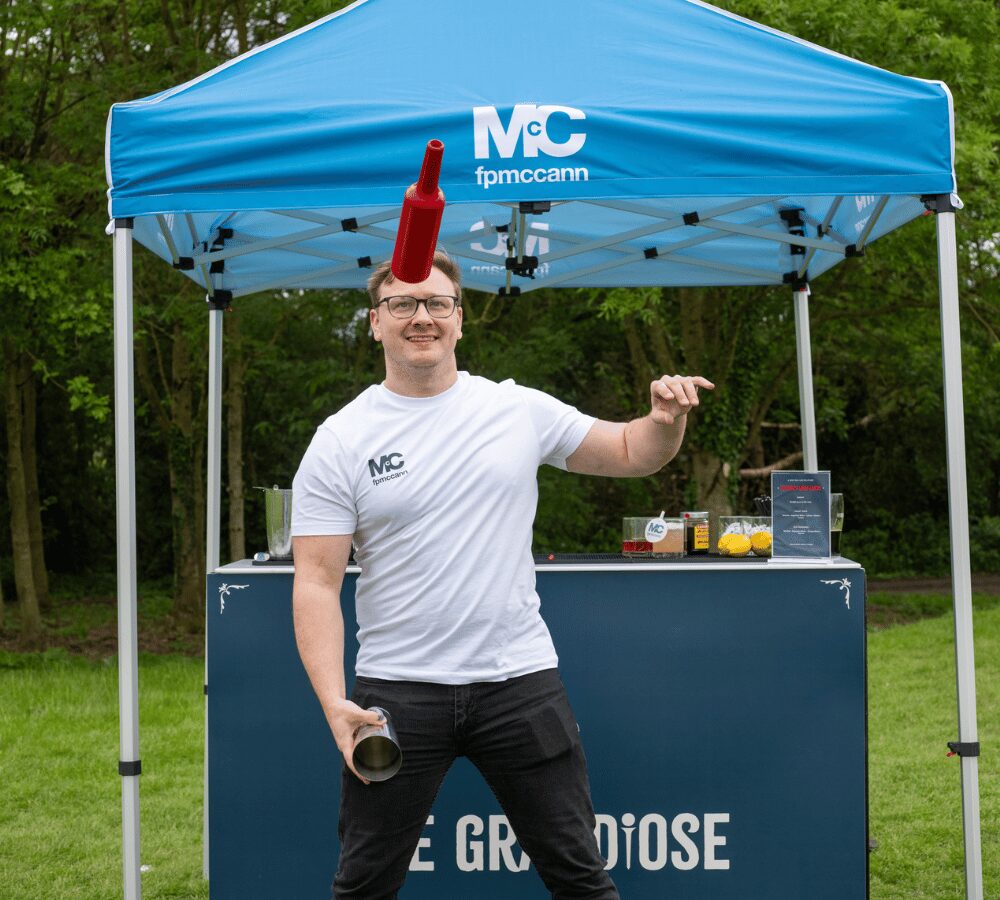 Participant juggling a bottle at an outdoor sponsored golf day beside a branded drinks station.